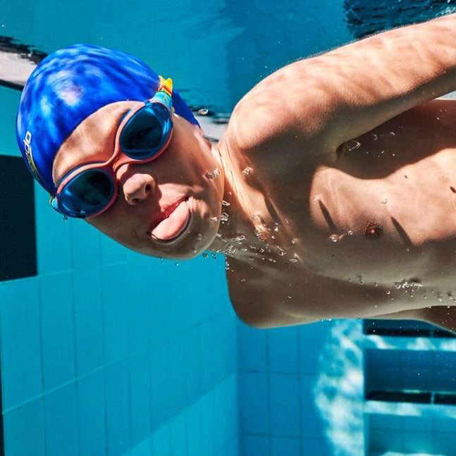 Junior swimmer wearing blue Speedo Biofuse 2.0 goggles with pink frames and a blue swim cap underwater.